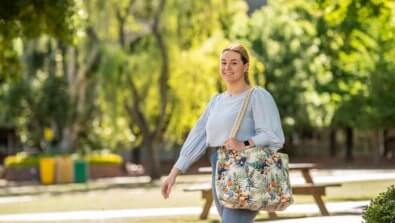A person walking outdoors with a floral bag, wearing a light blue blouse and jeans, in a park setting.