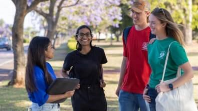 A group of four people standing outdoors, smiling and talking, with trees in the background.