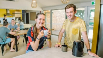 Two people smile at a kitchen counter with a kettle and cup. Others are sitting at a table in the background.