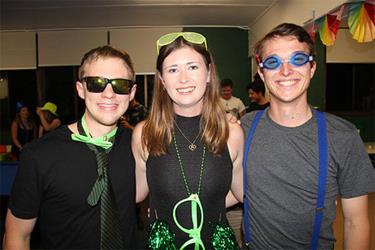 Three people smiling at a party, wearing festive attire and accessories such as sunglasses and novelty glasses.