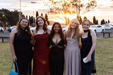 Five people standing in formal attire on a grassy area during sunset.
