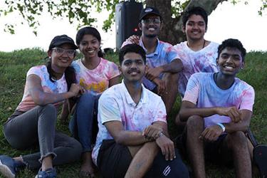 A group of six smiling young adults sitting on grass, wearing color-splattered clothes, likely during a Holi celebration.