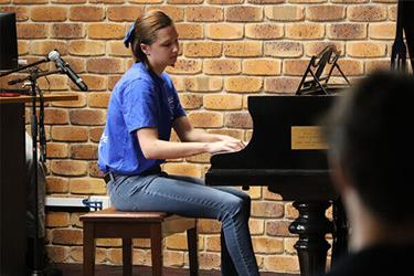 A person in a blue shirt plays a grand piano in front of a brick wall, with a microphone setup nearby.