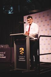 A person stands at a podium with a microphone, wearing a white shirt and red tie, at the University of Southern Queensland event.