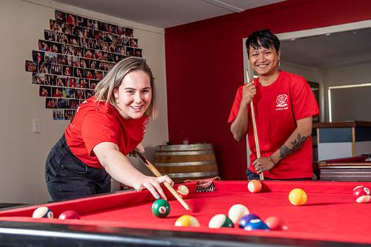 Two people in red shirts playing pool, with one person aiming the cue stick and the other smiling. A wall with photos is in the background.