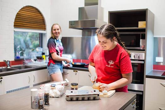 Two people are cooking in a kitchen. One is chopping vegetables, and the other is using a stove. Kitchen items like eggs and spices are on the counter.