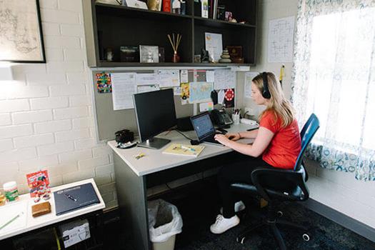 A person in a red shirt sits at a desk using a computer and wearing headphones. The workspace has a monitor, laptop, books, and items on shelves.