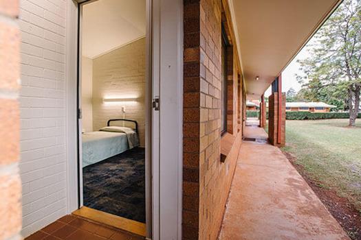 Exterior view of a brick building with an open door showing a bedroom inside. The room has a bed with a light above it. A grass lawn is visible to the right.