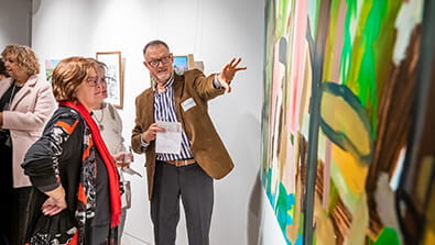 A man gestures toward a colorful painting in a gallery, conversing with a woman. Other people are viewing art in the background.