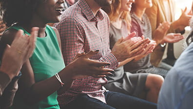 A group of people sit in a row, clapping their hands. Some wear casual clothing.