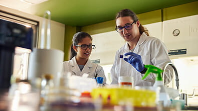Two scientists in a laboratory, wearing white coats and glasses, examine a small object. Various lab equipment is visible in the foreground.