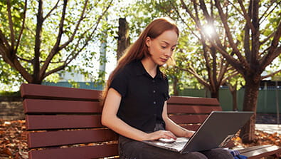 A person with long hair sits on a park bench using a laptop, surrounded by trees on a sunny day.