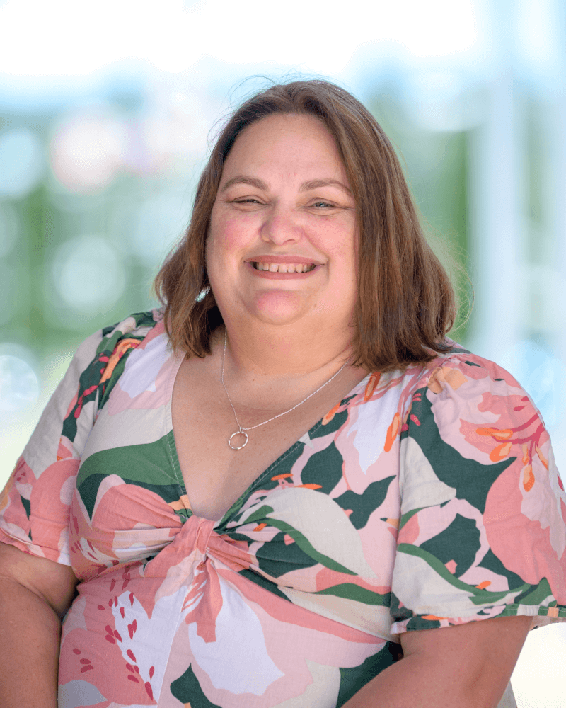 A woman with shoulder-length brown hair is smiling at the camera while wearing a floral-patterned dress and a silver necklace. The background is softly blurred.