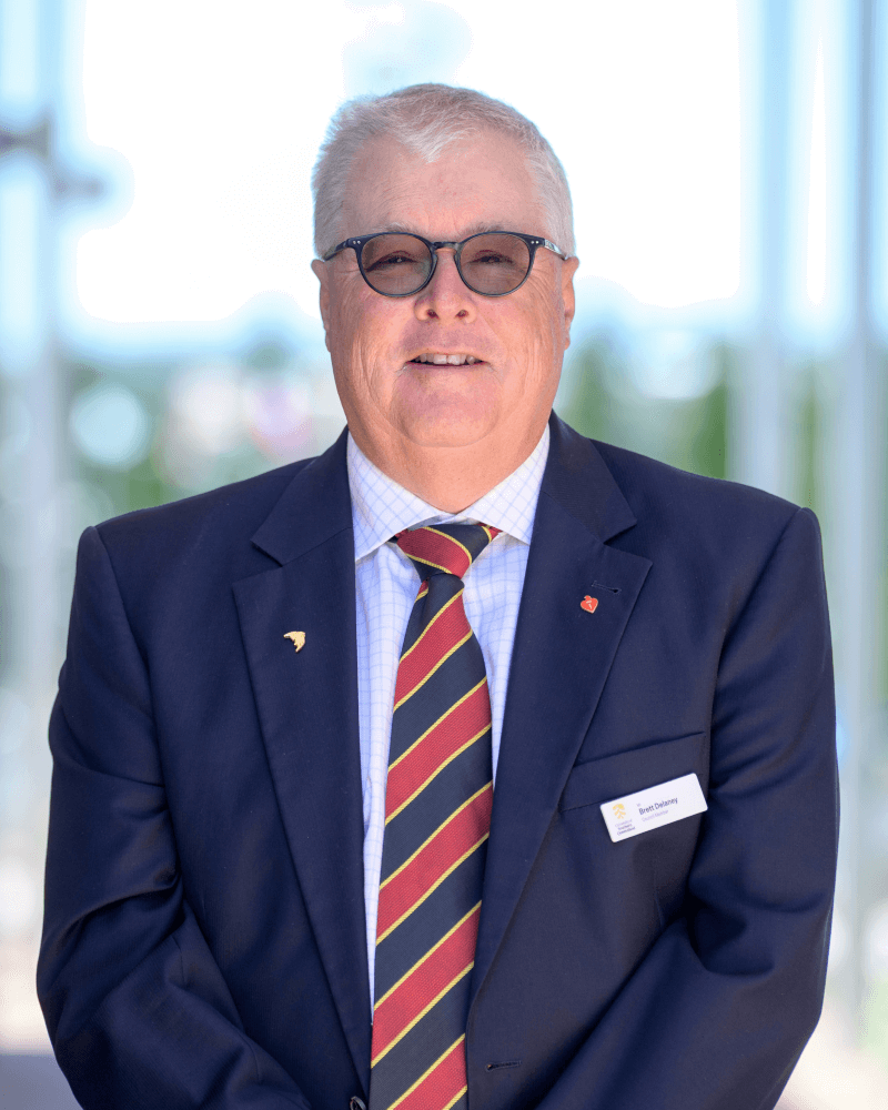 An older man with short gray hair and glasses wears a navy suit, striped tie, and name badge, standing outdoors in daylight.