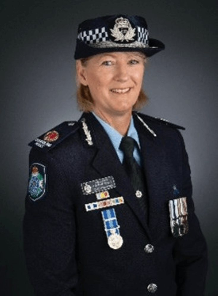 A police officer in formal uniform with medals and a checked hat, standing and smiling in front of a plain dark background.