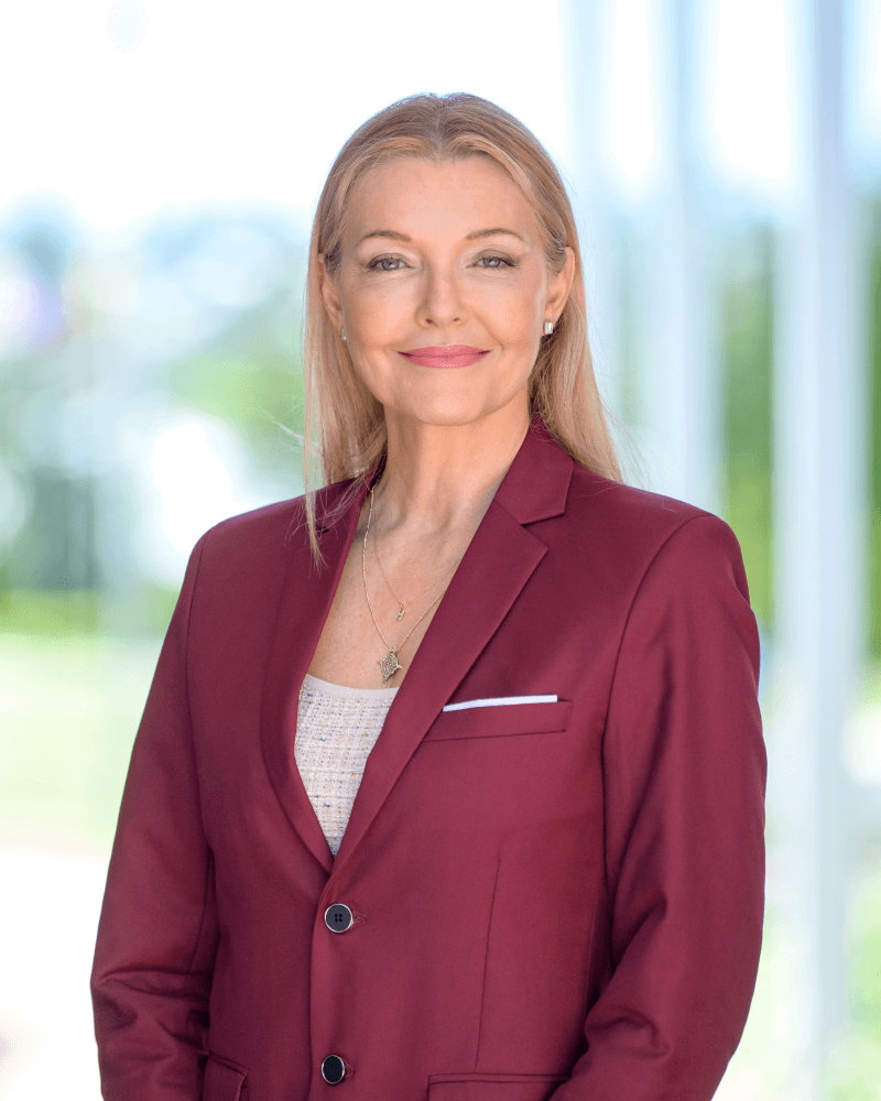 A woman with long blonde hair, wearing a maroon blazer and light top, stands outdoors smiling at the camera.