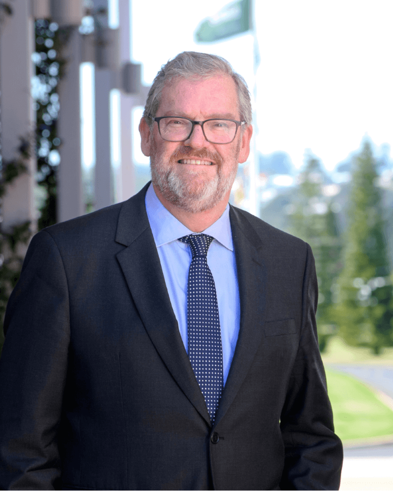 A man in a dark suit, blue shirt, and polka dot tie stands outside, smiling, with greenery and a blurred flag in the background.