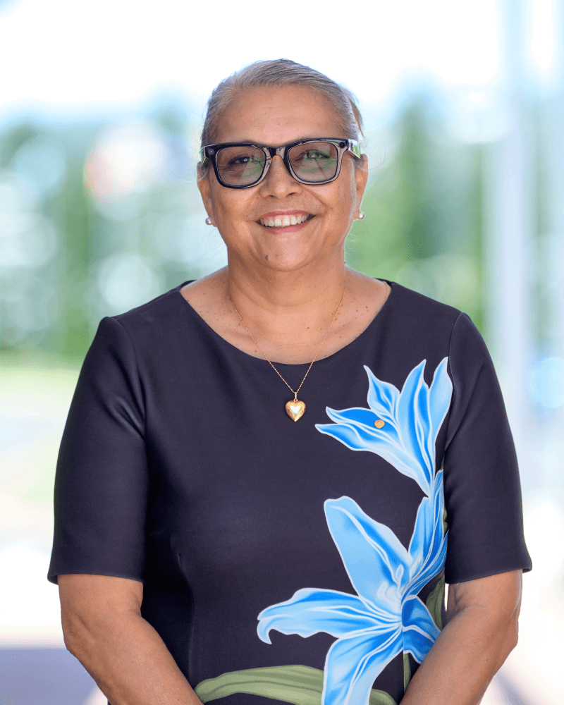 A woman with short gray hair, wearing glasses and a black dress with a large blue flower print, smiles while standing outdoors.