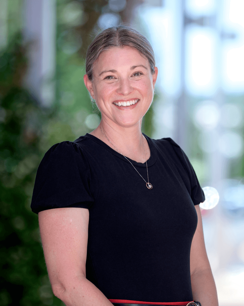 A woman with light brown hair tied back, wearing a black shirt and a necklace, smiles while standing outdoors with greenery and blurred buildings in the background.