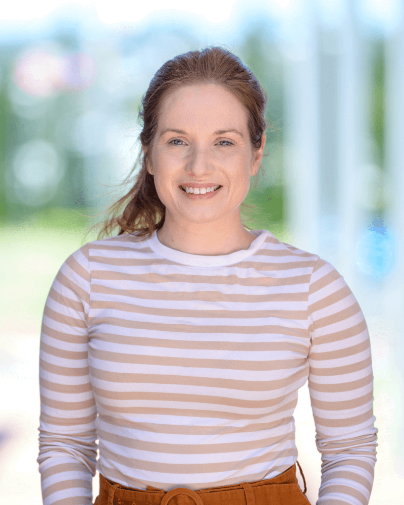A woman with light brown hair pulled back, wearing a beige and white striped long-sleeve shirt, stands outdoors and smiles at the camera.