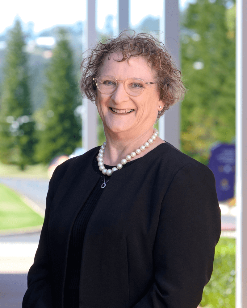 A smiling woman with short curly hair, glasses, and a pearl necklace stands outdoors in front of large windows and greenery.