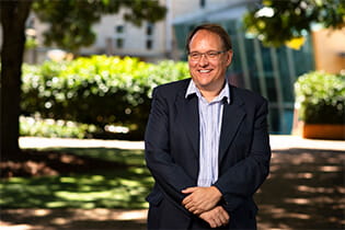 A man in a suit and glasses stands outdoors in a sunlit area with trees and a building in the background.