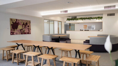 Modern office lounge with wooden tables, stools, cushioned seating booths, a front desk, wall art, and ceiling lights.