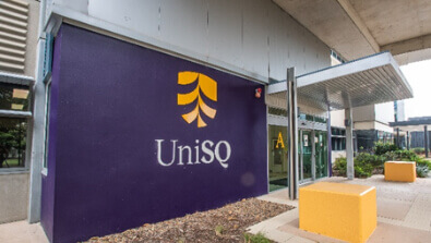 Exterior view of a building with a purple wall displaying the UniSQ logo and yellow seating blocks near the entrance.