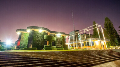 Building covered in greenery with illuminated edges, beside a modern structure with white frames, set against a purple evening sky and wide stone steps in the foreground.