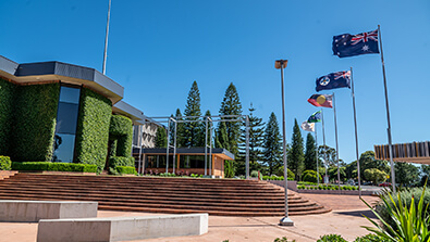A modern building with large windows and green ivy, surrounded by steps, trees, and five flags on flagpoles under a clear blue sky.