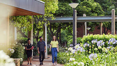Three people walk along a garden path beside a building, surrounded by green trees and blooming flowers on a sunny day.