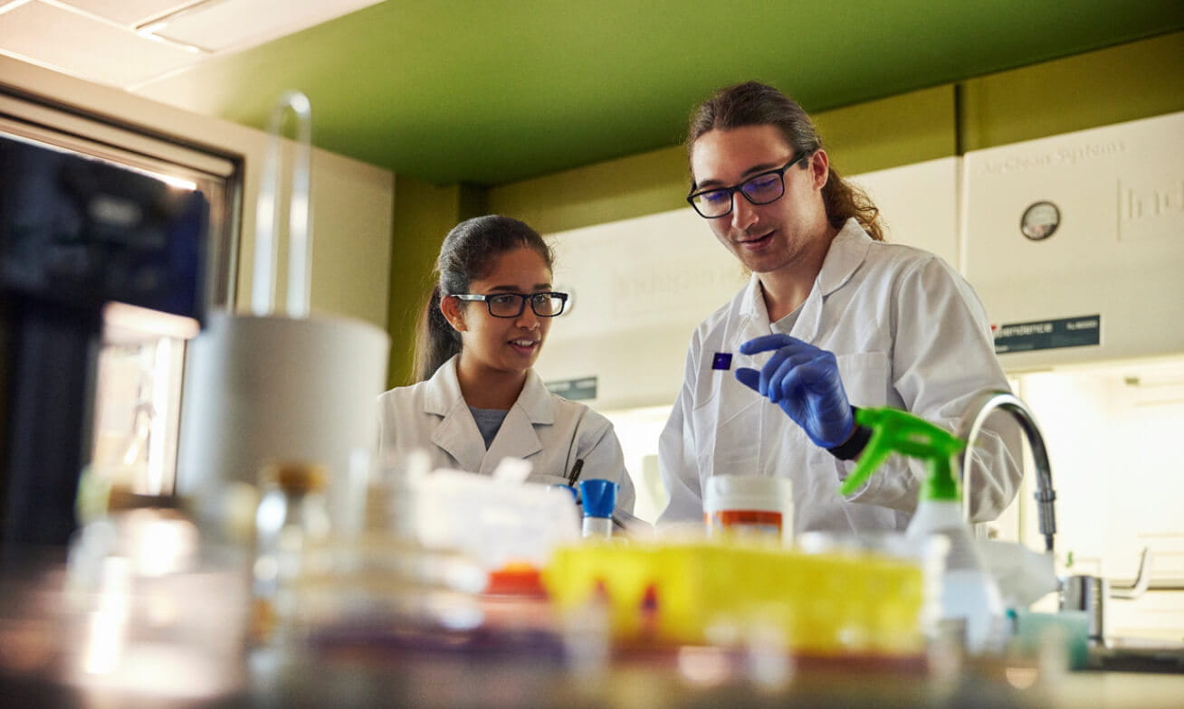 Two scientists wearing lab coats and safety glasses work together in a laboratory, examining a small sample slide among various equipment and containers.