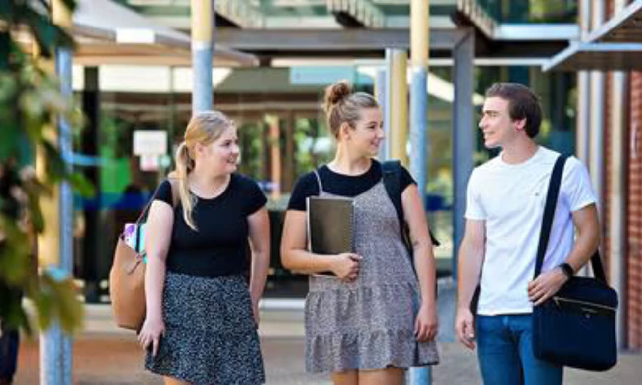 Three young adults walk outdoors near a building entrance, carrying bags and books. They are engaged in conversation and appear to be students.