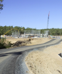 A construction site at UniSQ Springfield with scaffolding structures, cranes, and equipment is visible near a forested area, with a curved gravel road in the foreground.