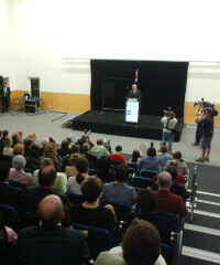 Former Australia Prime Minister John Howard stands at a podium on a stage addressing an audience in a lecture hall, with cameras and crew recording at UniSQ Springfield.