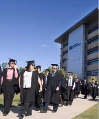 A group of graduates at UniSQ Springfield wearing caps and gowns walk along a path outside a modern academic building on a sunny day.