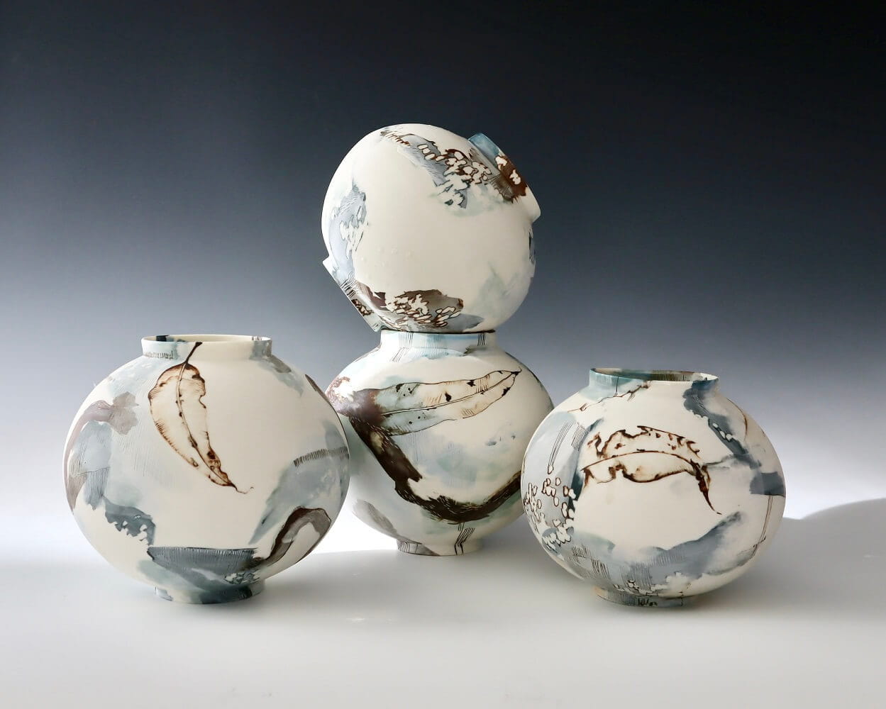 Four round, handcrafted ceramic vases with abstract blue, brown, and white patterns displayed on a flat surface against a neutral gradient background.