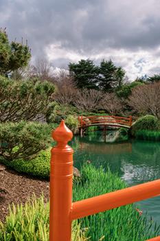 Orange rail and post in the foreground, with a small orange bridge crossing over a pond surrounded by lush greenery and cloudy sky in a landscaped garden.