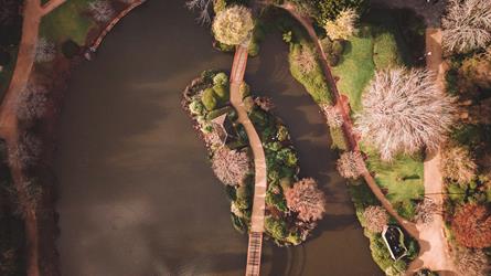 Aerial view of a winding wooden bridge over a pond, with gardens and sparse trees on either side, and a small gazebo on an island in the center at UniSQ Toowoomba Japanese Garden.