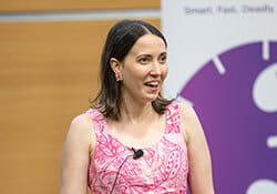 A woman in a pink patterned dress speaks at a podium with a microphone, standing in front of a blurred background and a purple and white sign.