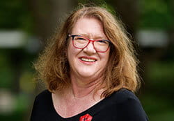 A woman with shoulder-length light brown hair and red glasses smiles at the camera, wearing a black top, outdoors with greenery in the background.