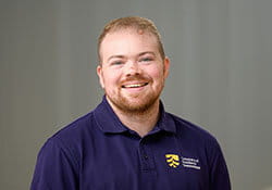 A man with short hair and a beard, wearing a navy blue polo shirt with a yellow and white logo, smiles at the camera against a plain background.