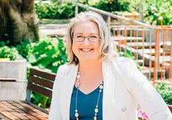 A woman with shoulder-length blonde hair and glasses is sitting on a bench outdoors, smiling. She is wearing a white blazer, a blue top, and a beaded necklace. Greenery is in the background.