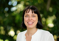 Woman with short dark hair and bangs, wearing a white blouse, smiling outdoors with green foliage in the background.