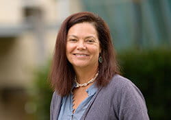 A woman with shoulder-length brown hair, wearing a gray cardigan and blue shirt, stands outdoors and smiles at the camera.