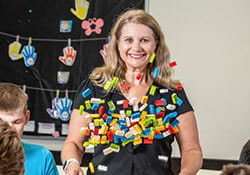 A smiling woman stands in a classroom with colorful plastic blocks stuck to her shirt, while students sit nearby.