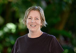 A woman with short blond hair, wearing a black t-shirt, stands outdoors in front of green foliage, smiling at the camera.