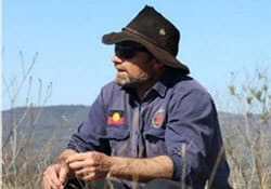 A man wearing a brown hat, sunglasses, and a blue shirt with an Aboriginal flag patch sits outdoors in a dry grassy area with hills in the background.