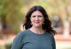 Woman with long brown hair wearing a green textured top stands outdoors with blurred trees and greenery in the background.