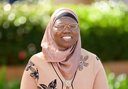 Woman wearing glasses and a light brown hijab smiles at the camera, standing outdoors with greenery in the blurred background.
